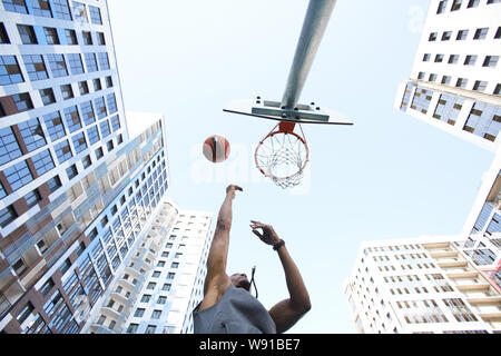 Low angle view at African basketball player shooting slam dunk against sky in urban background, copy space