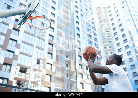 Portrait of sportive African-American man throwing ball at hoop while playing basketball in urban setting, copy space Stock Photo