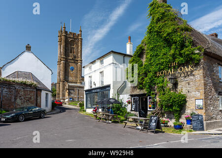 Cerne Abbas, England, UK - June 29, 2019: A man drinks outside The Royal Oak pub in the village of Cerne Abbas, with St Mary's Church behind. Stock Photo