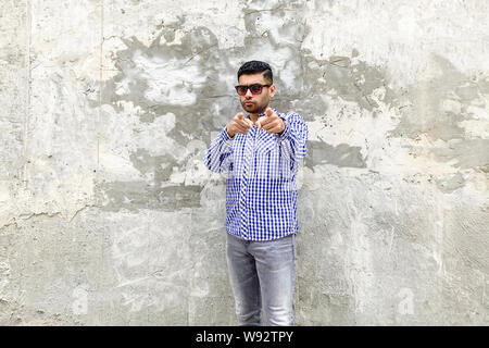 Hey you. Portrait of serious handsome bearded young man in checkered blue shirt and sunglasses standing against concrete gray wall, looking and pointi Stock Photo