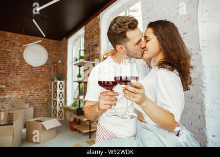 Young couple moved to a new house or apartment. Drinking red wine, caddling and relaxing after cleaning and unpacking. Look happy and confident. Family, moving, relations, first home concept. Stock Photo