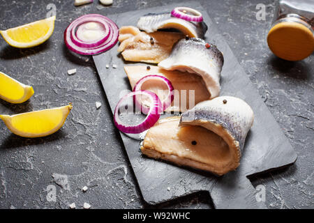 Herring fillet with salt, herbs, onion and lemon on black plate on stone background. Healthy food Stock Photo