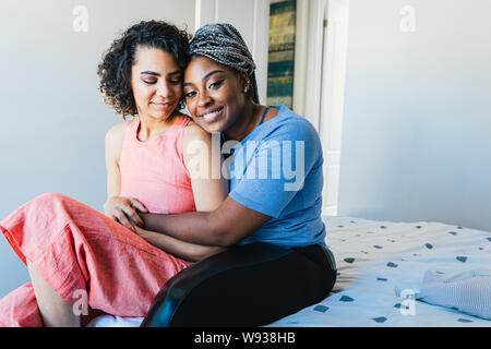 Portrait of cheerful woman embracing girlfriend while sitting on bed Stock Photo