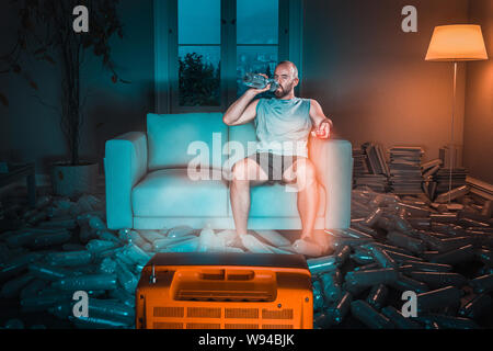 man watches tv on the sofa and drinks from a plastic water bottle, floor full of used bottles. Concept of exaggerated use of plastic and recycling. Stock Photo