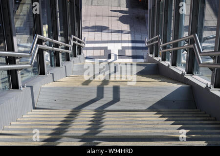 sunlight casting shadows on concrete steps leading from pedestrian bridge over railway station to platform Stock Photo