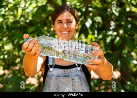 A young woman holding up a plastic bottle refilled with clear natural spring water. orange trees and green leaves behind Stock Photo