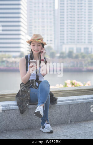 Beautiful asian solo tourist woman relaxing and enjoying listening the music on a smartphone in urban city downtown. Vacation travel in summer. Stock Photo