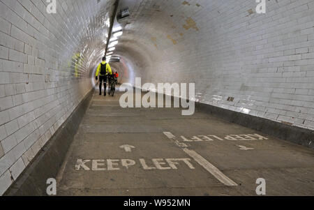 Cyclist wearing yellow jacket walking with his bicycle inside Greenwich foot tunnel under river Thames, more blurred pedestrians in background, keep Stock Photo