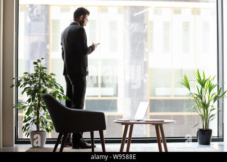 Confident businessman standing near window, using phone Stock Photo