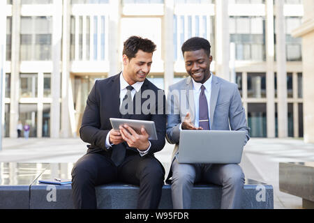 Two businessmen sitting outdoors talking to each other and working on digital tablet and laptop computer Stock Photo