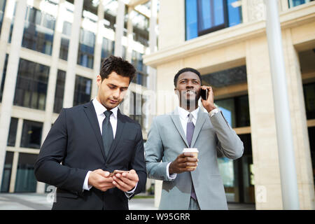 Two young businessmen in suits using mobile phones typing a message and have a conversation while walking along the street in the city Stock Photo