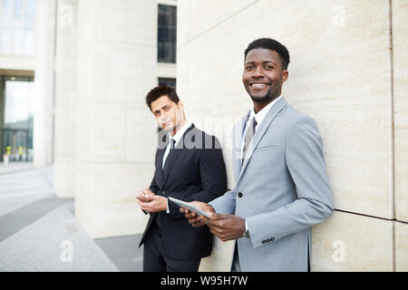 Portrait of African young office worker standing in suit using tablet pc and smiling at camera with his colleague standing in the background in the city Stock Photo