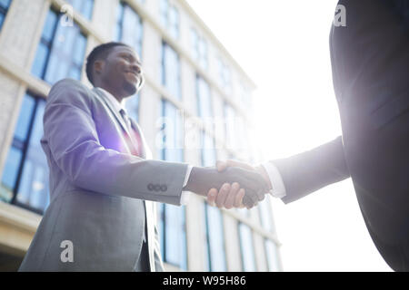 African young businessman in suit smiling and shaking hands to his partner while they standing outdoors Stock Photo