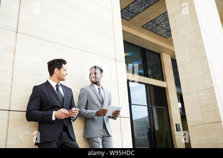 Two young business colleagues wearing formalwear leaning on the wall of office building and using modern gadgets outdoors Stock Photo
