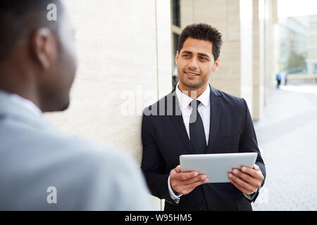 Confident office worker in suit working on digital tablet while talking to his partner in the city outdoors Stock Photo