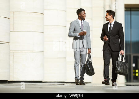 Two multiethnic young businessmen in formal clothing walking along the street after work day and discussing some moments in the city Stock Photo
