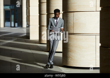 Young African businessman wearing suit leaning on the column of office building reading a document and drinking coffee outdoors Stock Photo