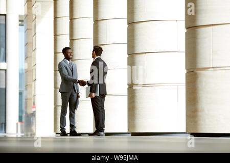 Two business partners in suits standing and shaking hands during their meeting outdoors near the office building Stock Photo
