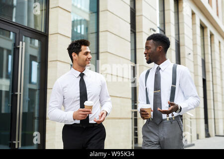 Two young multiethnic businessmen discussing something and drinking coffee while walking along the street in the city Stock Photo