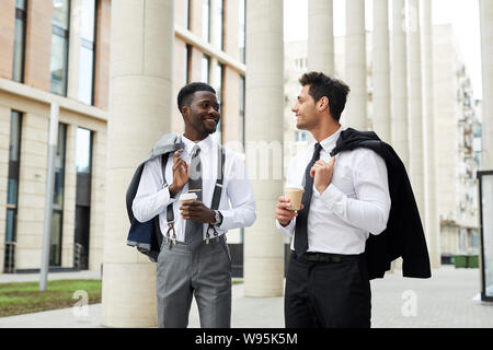 Two multiethnic office workers smiling and talking to each other while walking along the street and drinking coffee from disposable cups Stock Photo