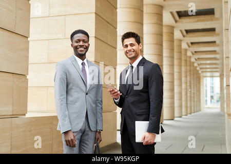 Portrait of two successful multiethnic businessmen in formal clothing smiling at camera while standing outdoors near the modern building Stock Photo