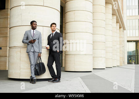 Two young managers in formal clothing with mobile phone and coffee drink looking away while standing near the office building outdoors Stock Photo