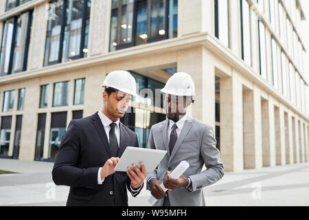 Two young multiethnic engineers wearing hardhats standing in the city and using digital tablet together Stock Photo