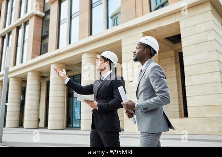 Two multiethnic engineers wearing suits and hardhats looking at new building while standing outdoors in the city Stock Photo