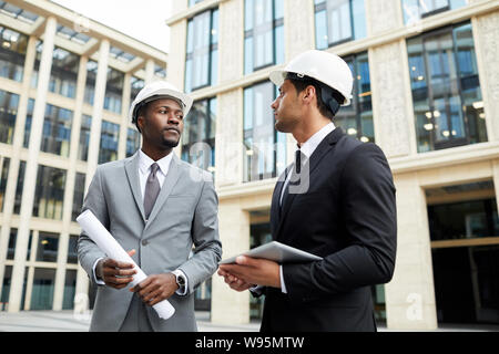 Two multiethnic engineers in hardhats discussing new project and working in team while standing in big city among modern buildings Stock Photo