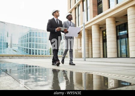 Two multiethnic architects in formal clothing and hardhats holding blueprint walking along the street and looking at modern building Stock Photo