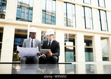 Two multiethnic businessmen sitting together in the city examining blueprint and using digital tablet in online work Stock Photo