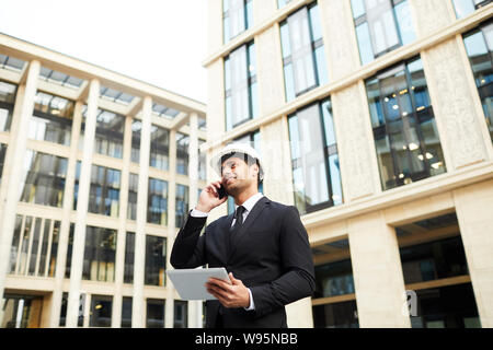 Young architect in formal wear and hardhat holding tablet pc talking on mobile phone and smiling while standing in the city Stock Photo