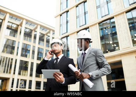 Young engineer in suit and helmet talking on mobile phone with his colleague standing near by they working in the city Stock Photo