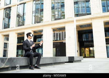 Young businessman wearing virtual reality goggles has online communication while sitting outdoors near the office building Stock Photo