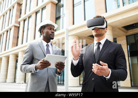 Young businessman wearing virtual reality glasses standing outdoors together with his colleague who using digital tablet in the city Stock Photo