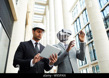 Serious young businessman using digital tablet and controlling the process of virtual reality game of his colleague in the city Stock Photo