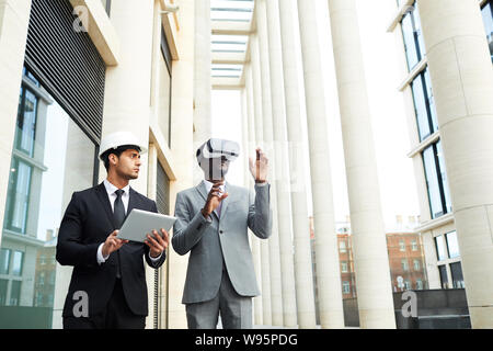 Young businessman in hardhat and suit using tablet pc and looking at his colleague while he wearing virtual reality goggles and walking in the city Stock Photo