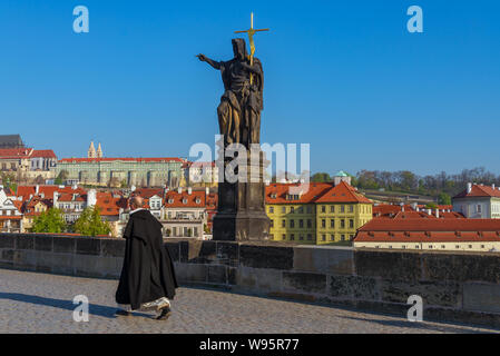 Bishop or priest walk near statue of Saint John the Baptist on pedestal and balustrades of Charles Bridge over Vltava river  in sunny day.