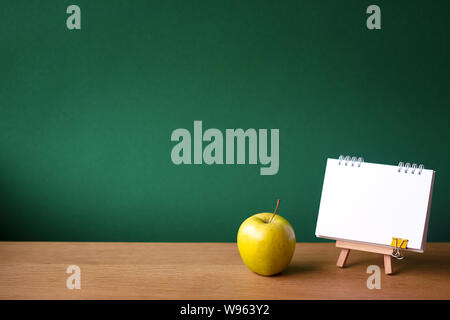 Back to school concept, open notebook on miniature easel and green apple on wooden surface on the backdrop of a clean green chalk board, selective foc Stock Photo