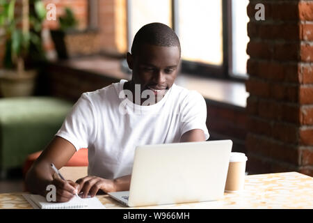 Smiling black male studying on laptop making notes in notebook Stock Photo