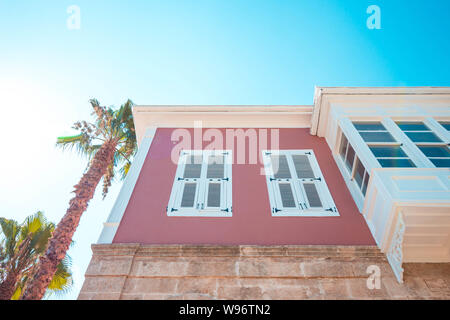 A palm tree next to the house is pink. The house has two large white windows and a balcony. House and palm tree against a clear blue sky. Stock Photo