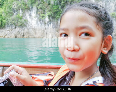 The little girl holding a camera and sitting in a tourist boat in the sea with the mountain. Close up kid wear life jacket smiling and looking. Stock Photo