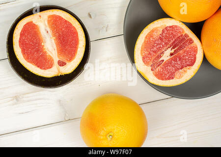 Group of three whole two halves of fresh pink grapefruit in dark ceramic bowl on gray ceramic plate flatlay on white wood Stock Photo