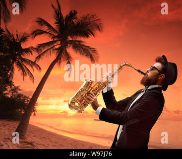 Male artist playing saxophone with a beach and a palm on sunset as a background Stock Photo