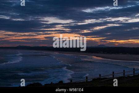 Beach from the headland at dusk, almost dark, a sea of clouds above and an ocean of water below, orange sky behind the grey, waves breaking near beach Stock Photo