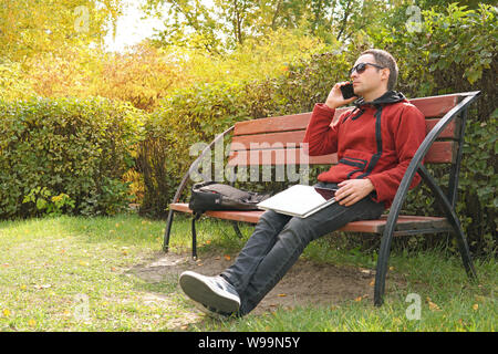 Happy casual man with laptop and take away coffee talking on mobile. Handsome male freelancer working outdoors in the park. Technology, communication Stock Photo