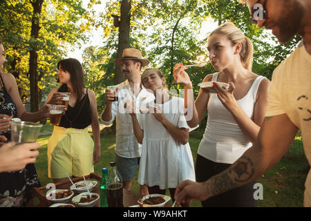 Group of happy friends eating and drinking beers at barbecue dinner on sunset time. Having meal together outdoor in a forest glade. Celebrating and relaxing. Summer lifestyle, food, friendship concept. Stock Photo