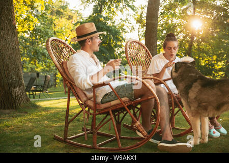 Happy couple eating and drinking beers at barbecue dinner on sunset time. Having meal together outdoor in a forest glade. Celebrating and relaxing. Summer lifestyle, food, family, relation concept. Stock Photo