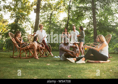 Group of happy friends eating and drinking beers at barbecue dinner on sunset time. Having meal together outdoor in a forest glade. Celebrating and relaxing. Summer lifestyle, food, friendship concept. Stock Photo