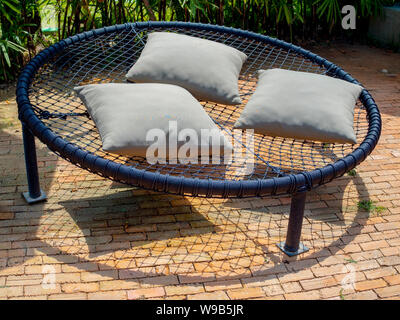 Black round macrame sofa bed on multiple ropes with three pillows on brick floor in garden. Stock Photo